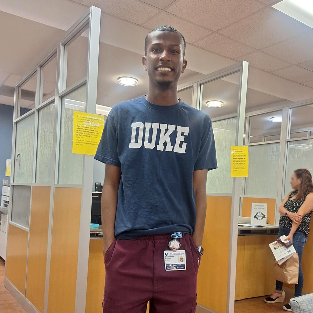 A young man in a medical office wearing scrub pants, a work badge, and a dark blue T-shirt reading 'DUKE.'
