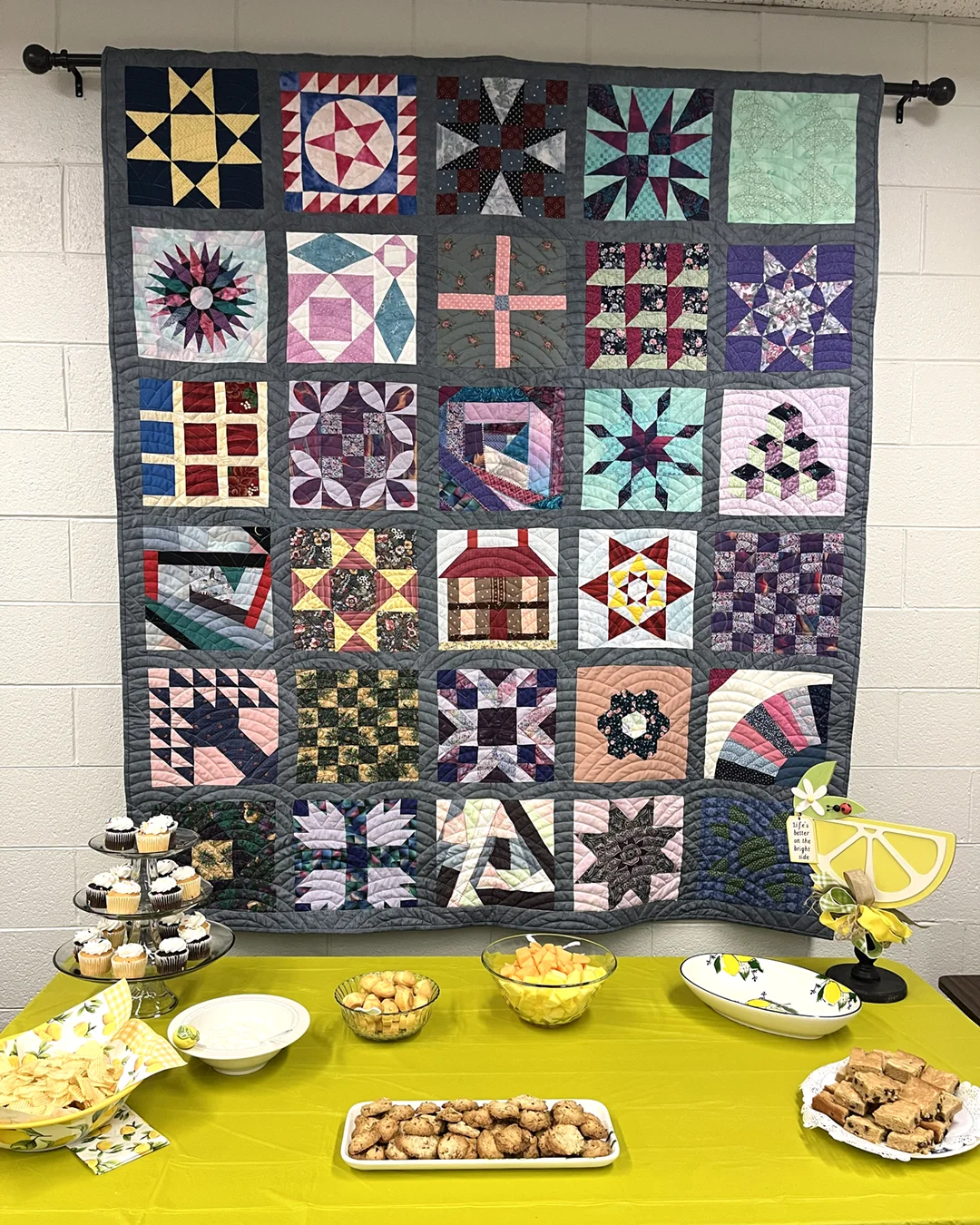 A patchwork quilt hanging on a wall, comprised of 30 eclectic patchwork squares (5x6) each displaying a different quilting pattern or technique. A table of refreshments stands below.