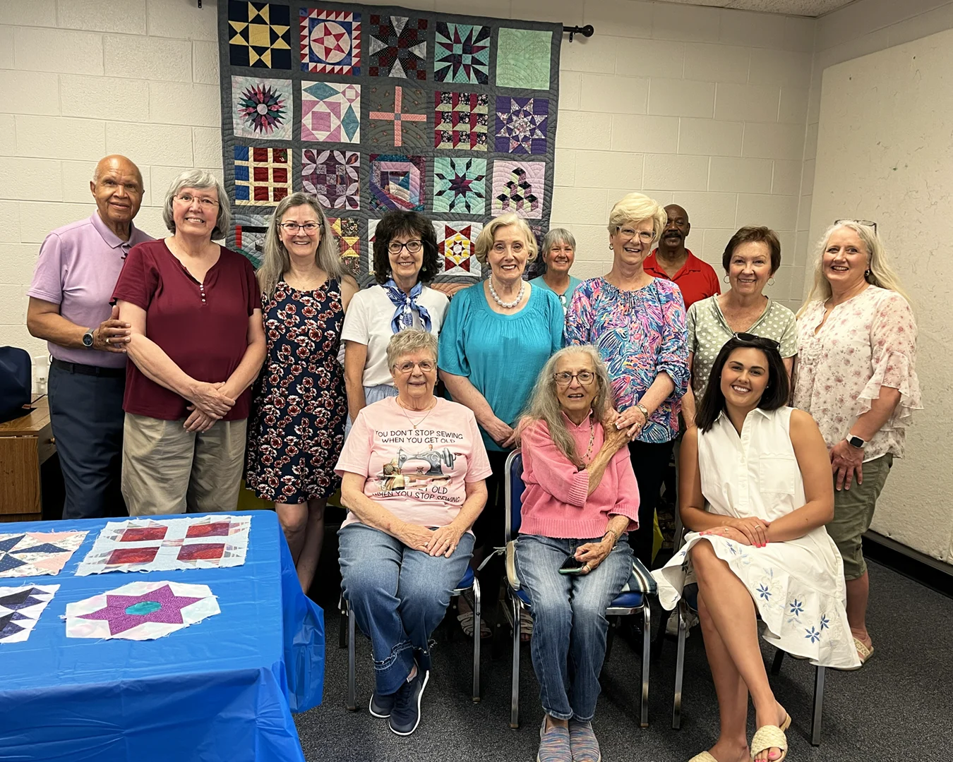 Thirteen adults pose in front of a patchwork quilt hanging on a wall.