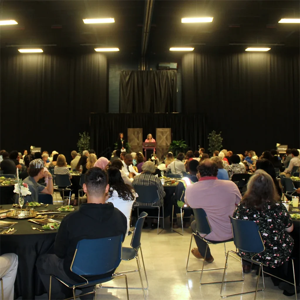 People seated at round banquet tables in a large room listen to a woman speaking on a stage.