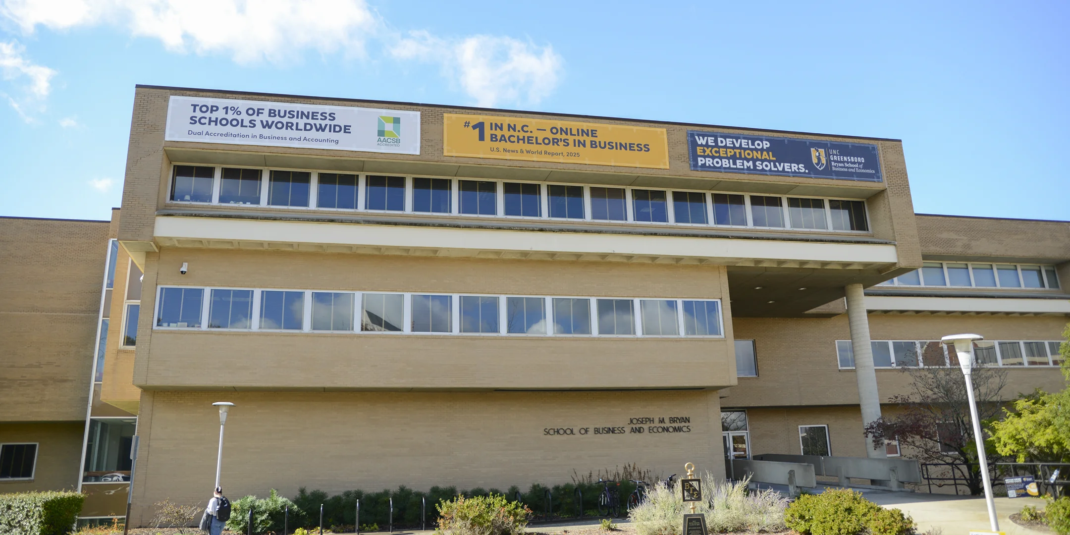 Wide shot of a three-story brick building labeled 'Joseph M Bryan School of Business and Economics,' with three vinyl banners showcasing UNCG accolades.