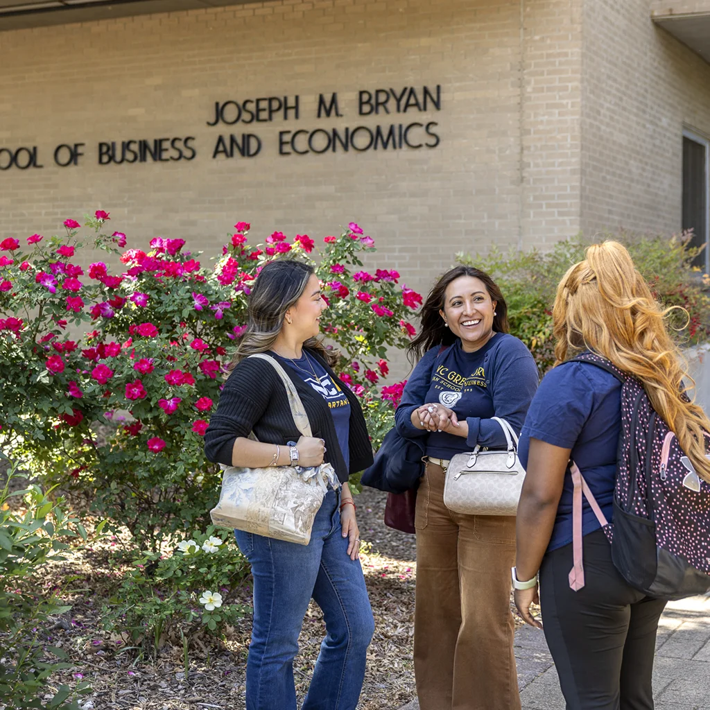 Three female college students wearing dark blue UNCG T-shirts converse in front of a brick building labeled 'Joseph M Bryan School of Business and Economics.'