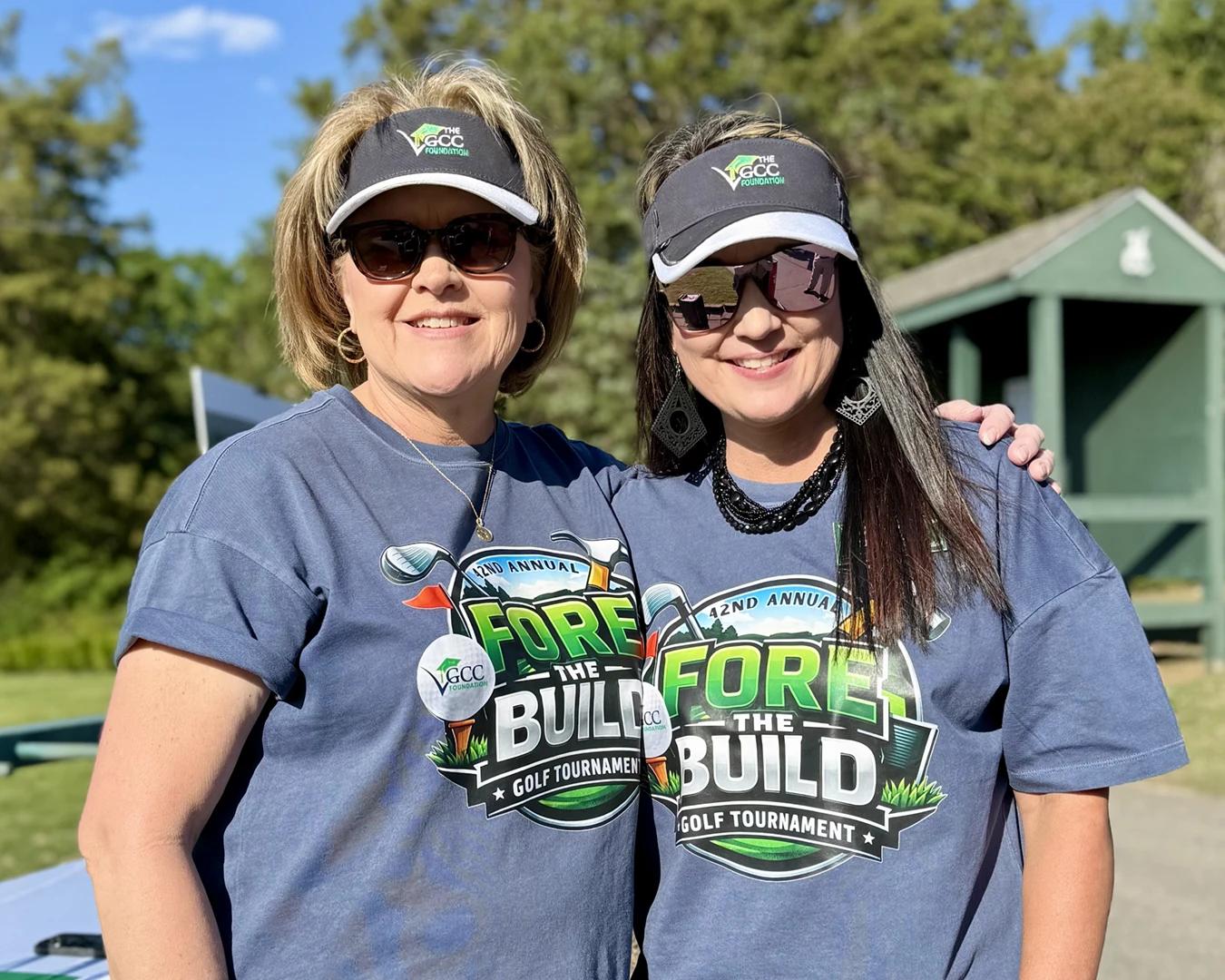 Two women stand outdoors with their arms around each other, while wearing matching golf tournament T-shirts and VGCCF visors.