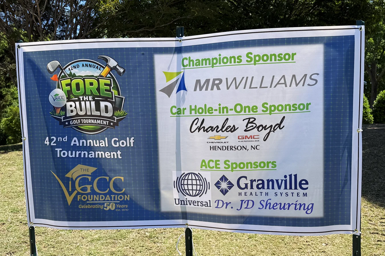 Outdoor banner displaying VGCCF 'Fore the Build' golf tournament sponsors. Champions Sponsor: MR Williams. Car Hole-In-One Sponsor: Charles Boyd Chevrolet Cadillac GMC. Ace Sponsors: Granville Health System, Dr. JD Sheuring, Universal Leaf.
