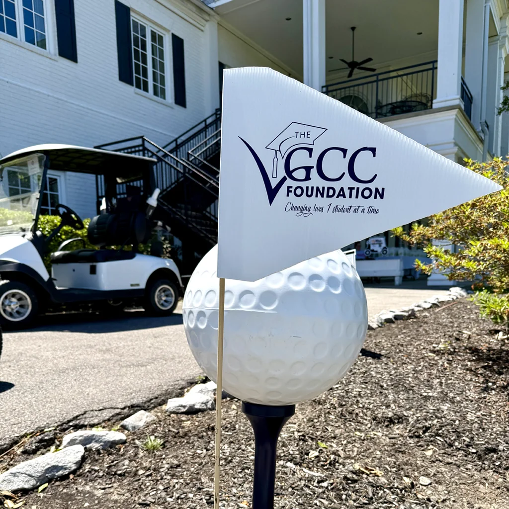 Promotional golf flag for The VGCC Foundation with tagline 'Changing lives 1 student at a time.' A large decorative golf ball, two golf carts, and the main building of Henderson Country Club are in the background.