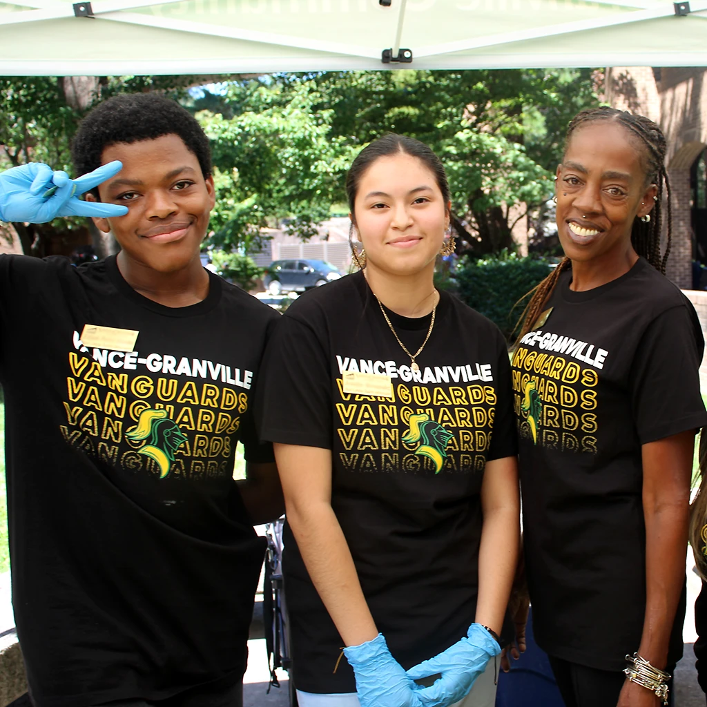 Three diverse college students wearing black T-shirts reading 'Vance-Granville Vanguards' while wearing blue rubber gloves to serve food outdoors.