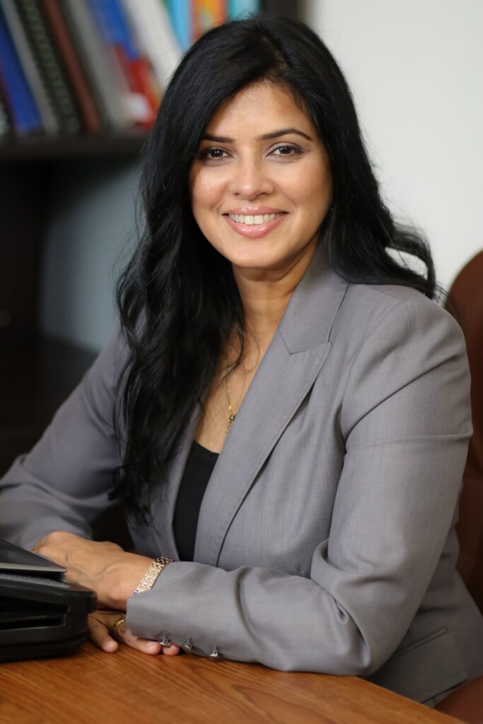 Professional headshot of a woman with long dark hair and a gray blazer, sitting at a wooden desk in an office.