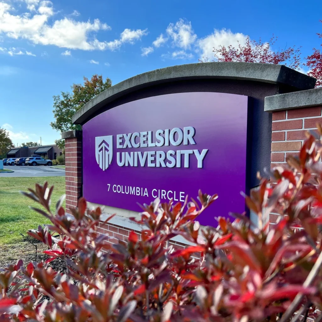 Closeup photo of a purple Excelsior University campus sign under a bright blue sky.