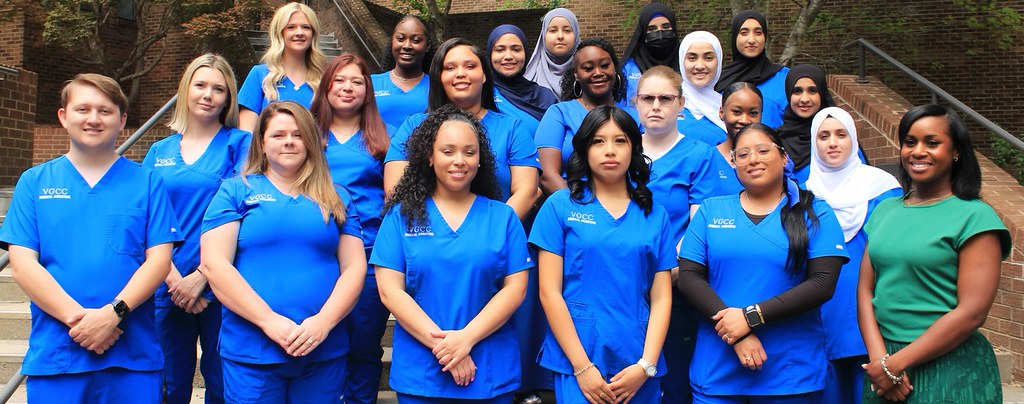 20 healthcare students in blue scrubs pose with their instructor for a class portrait.