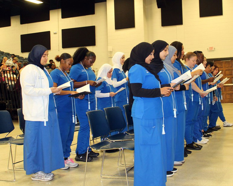 Two rows of healthcare students in blue scrubs stand and read aloud from a paper handout in front of a crowd.