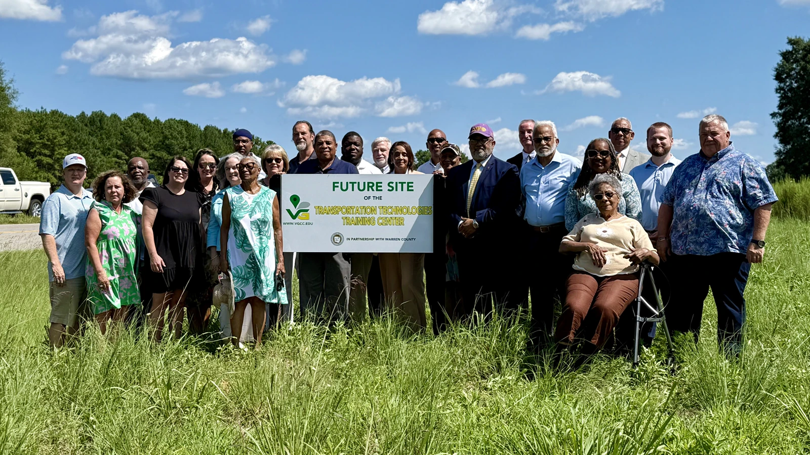 24 adults in business attire stand in a grassy field, surrounding a mounted sign reading 'Future Site of the Transportation Technologies Training Center, in partnership with Warren County.'