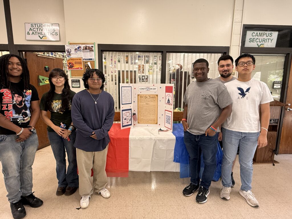 Six diverse students standing with a constitution tri-fold presentation in the student lounge on Main Campus.