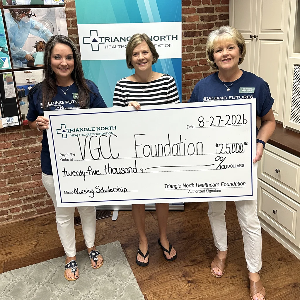 Three women stand in a building lobby, holding a large $25,000 check from Triangle North Healthcare Foundation to VGCC Foundation for nursing scholarships.