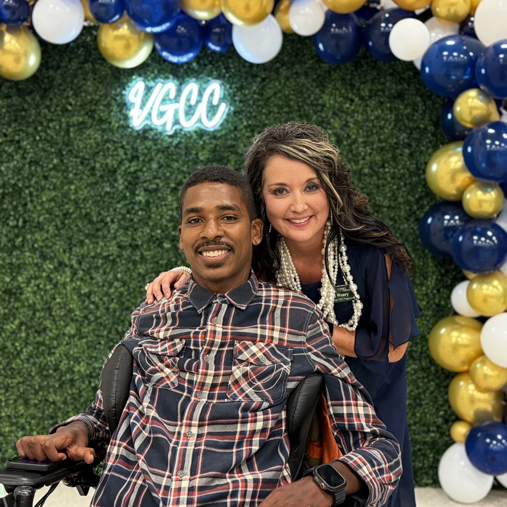 A woman stands with her arm around a young man in a motorized wheelchair; a stylized backdrop with balloons and a 'VGCC' neon sign in the background.