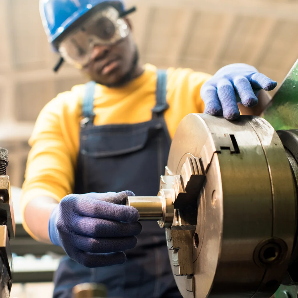 Male-presenting machine operator wearing safety goggles and hardhat adjusting equipment while working at production department of modern plant.