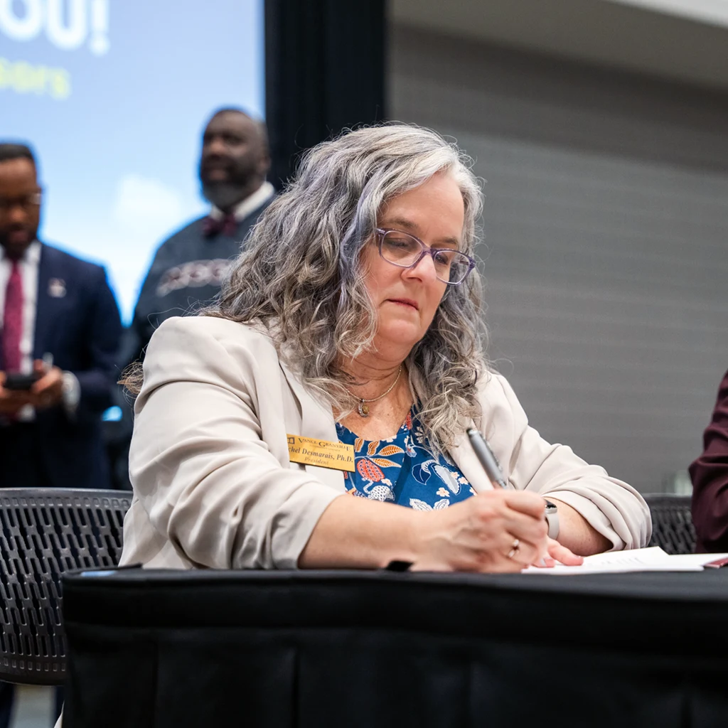 A middle-aged woman seated at a table uses a pen to sign a sheet of paper.