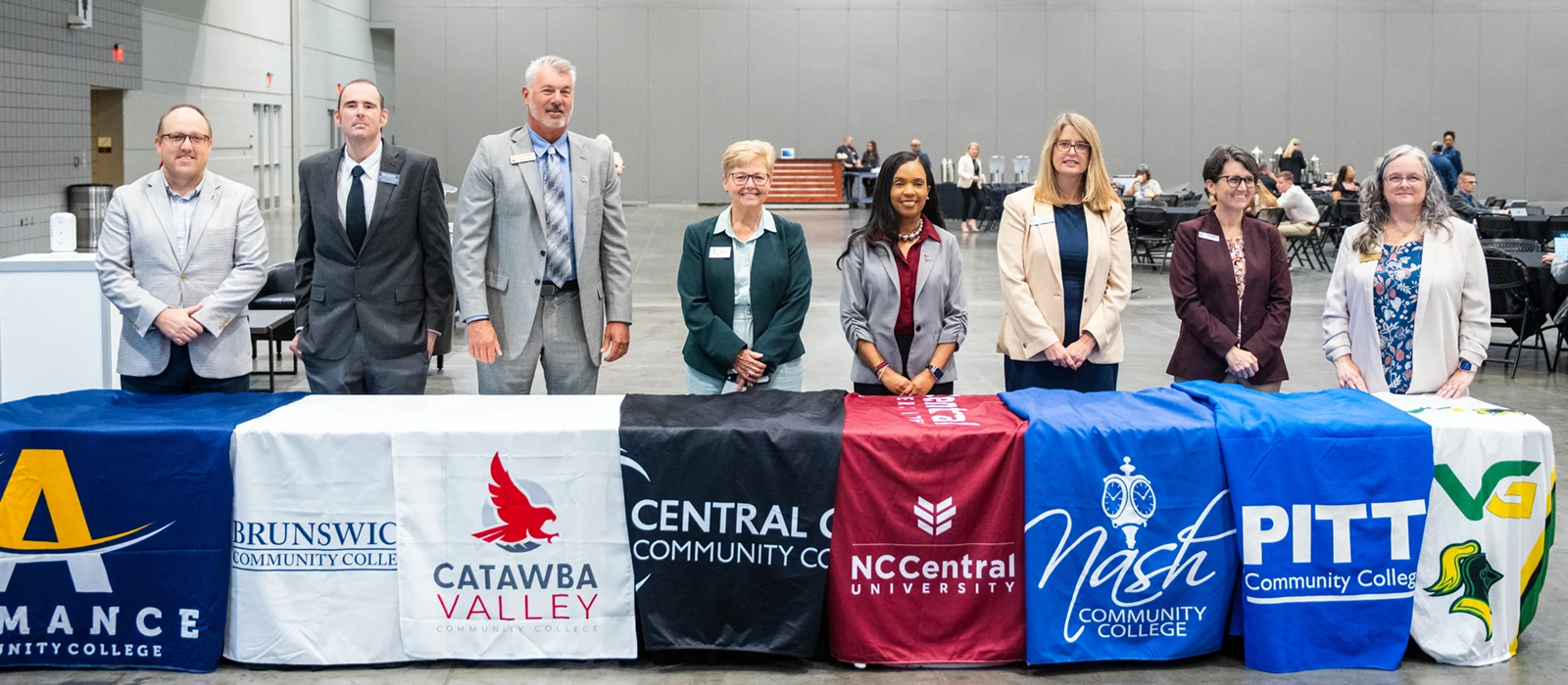 Eight adults in business attire stand behind a long table, which is covered in banners representing their respective colleges and universities.