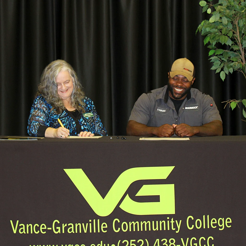 A middle-aged woman in business attire and a young man in a tradesman uniform both smile as they sit behind a table and sign papers.