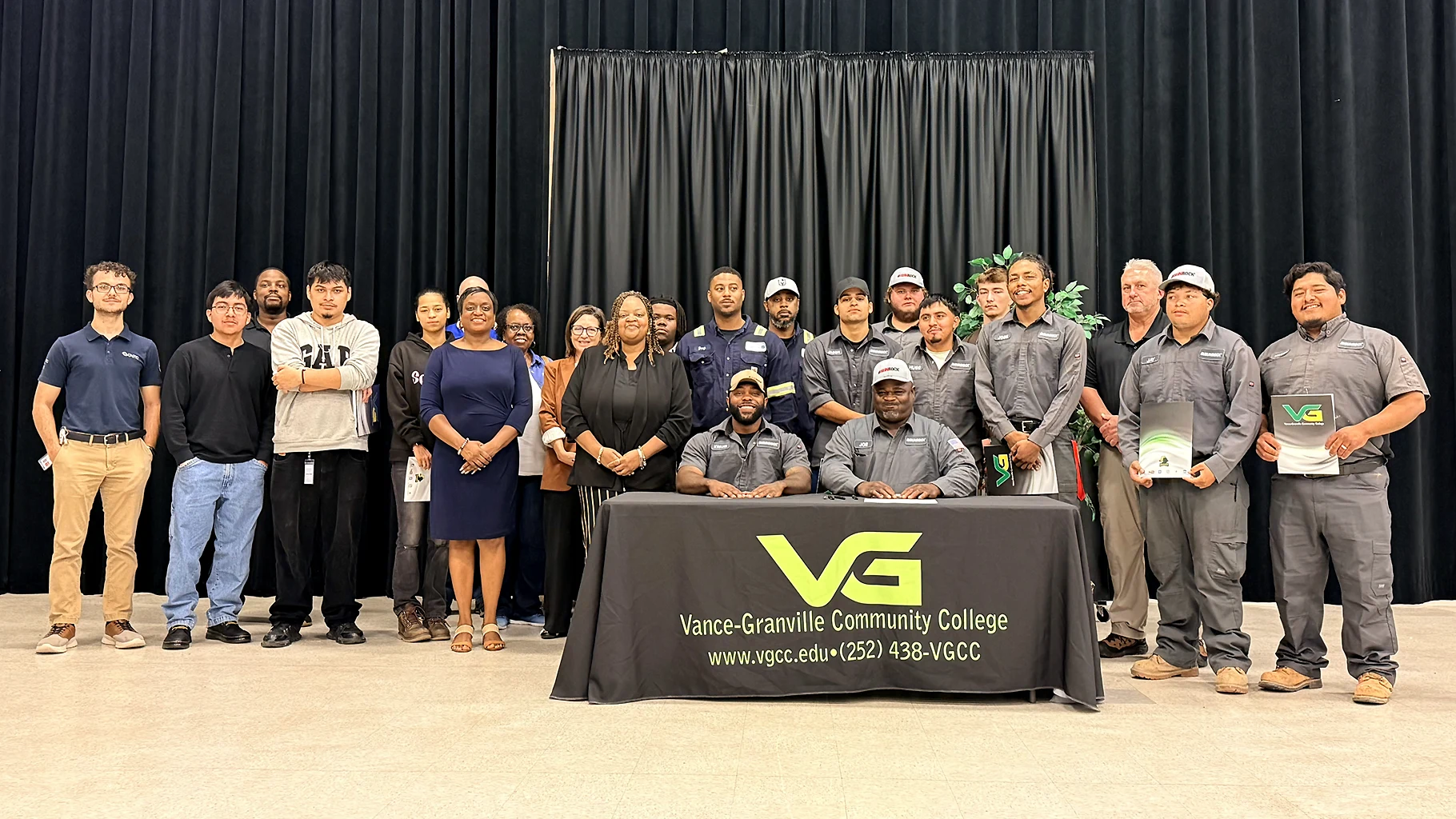 A diverse group of 23 adults gather behind a table covered in a cloth bearing the Vance-Granville Community College logo.