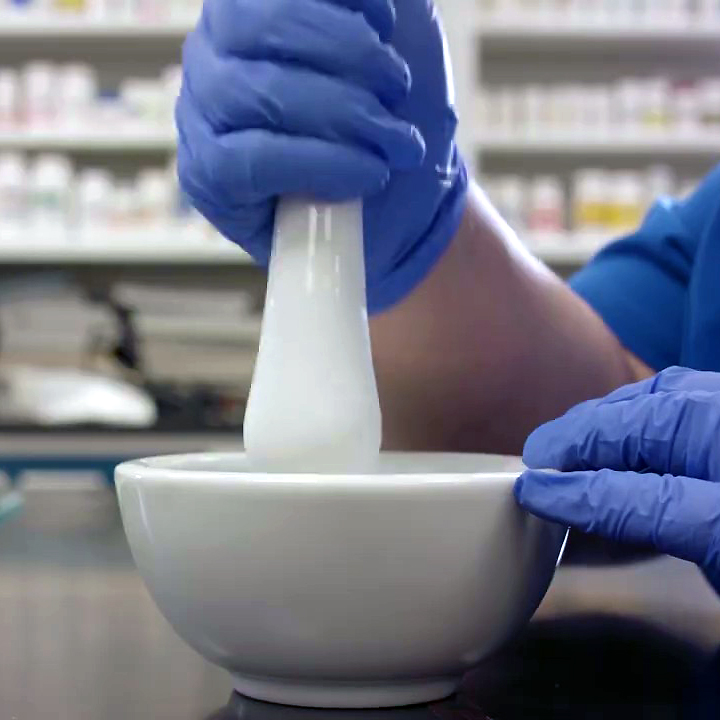 Close-up of hands wearing blue gloves while using a mortal and pestle, shelves of white bottles blurred in the background.